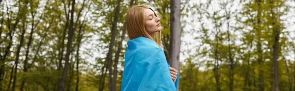 Вдохновлённая блондинка, держащая украинский флаг над своим телом в лесу с закрытыми глазами, баннер — стоковое фото