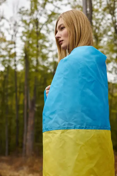 Side view of blonde young woman wrapping the Ukrainian flag over her body in forest scenery — Stock Photo