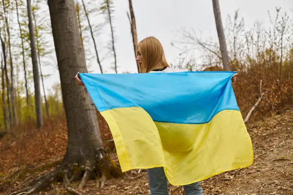 Back view of female young blonde hiker in sweater holding national Ukrainian flag in forest, freedom — Stock Photo