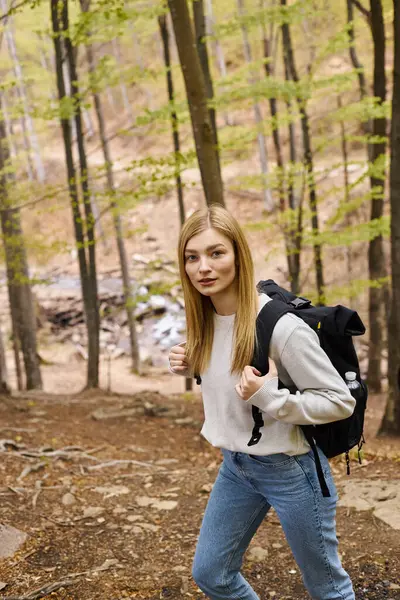 Neugierige blonde Wanderin mit Reiserucksack schlängelt sich durch den Wald und blickt in die Kamera — Stockfoto