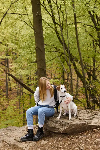 Liebevolle blonde Wanderin, in Jeans und kuscheligem Pullover, streichelt ihren treuen Hund, der im Wald sitzt — Stockfoto