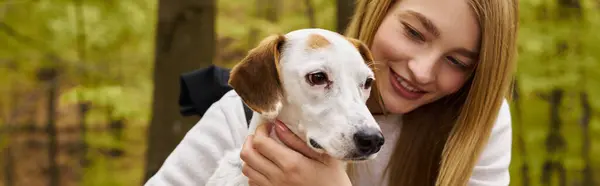 Femme blonde souriante étreignant son chien, prenant soin de son animal de compagnie tout en s'arrêtant en voyage en forêt, bannière — Photo de stock