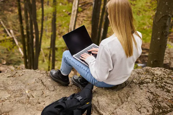 Vista trasera de la mujer senderista rubia sentada en la roca en el bosque con el ordenador portátil en sus rodillas con mochila - foto de stock