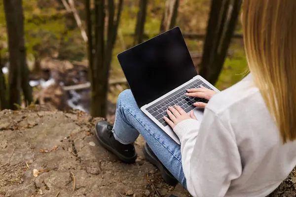 Vista trasera de una mujer rubia sentada en una gran roca en el bosque, utilizando un ordenador portátil para el trabajo remoto - foto de stock