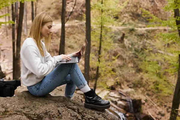Mujer rubia aventurera senderismo en el bosque, sentado en la roca con el ordenador portátil y la mochila - foto de stock