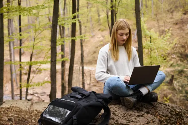 Young blonde woman with backpack hiking in woods, sitting on boulder with laptop and working — Stock Photo