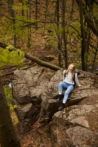 Young woman hiker with backpack sitting at halt on rocky cliff in forest looking in the sky — Stock Photo