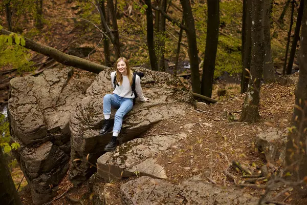 Улыбающаяся женщина-туристка с рюкзаком, сидящая на скалистой скале в лесу и смотрящая в камеру — стоковое фото