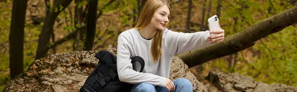 Mujer joven rubia tomando selfie sentado en la roca en alto en el bosque mientras caminaba, pancarta - foto de stock