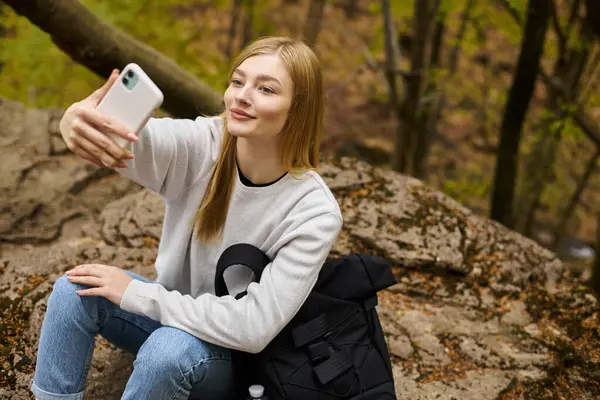 Sonriendo tranquila joven rubia tomando selfie mientras se relaja en el bosque mientras camina - foto de stock