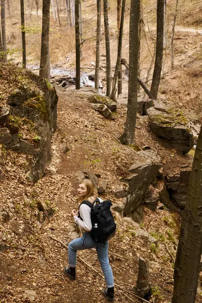 Back view of cheerful blonde female hiker wearing backpack crossing the forest with stream — Stock Photo