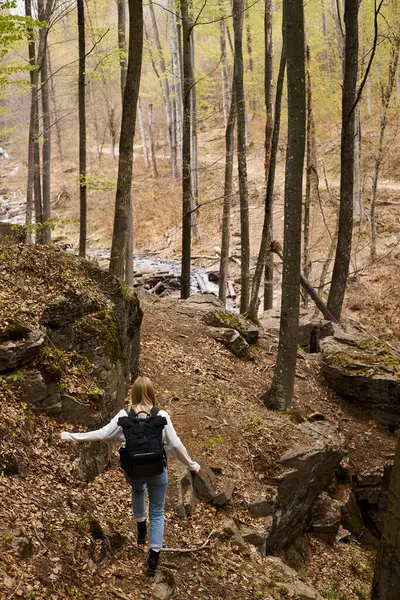 Vue arrière d'une jeune voyageuse blonde portant un sac à dos courant vers un ruisseau forestier — Photo de stock