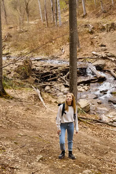 Jolie randonneuse blonde avec sac à dos de voyage, marchant près d'un ruisseau forestier regardant dans le ciel — Photo de stock