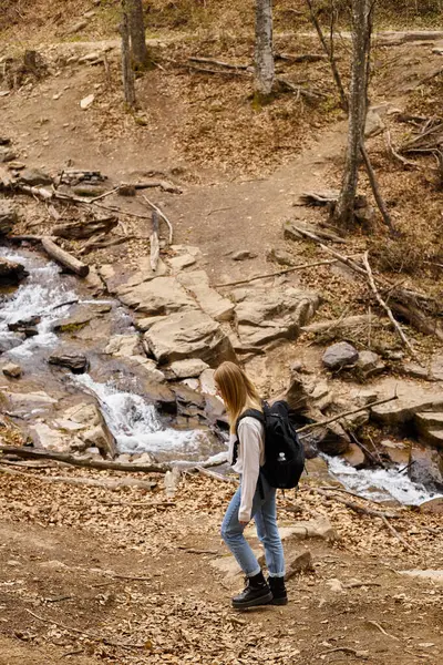 Young blond trekker with travel backpack, walking near forest stream enjoying views — Stock Photo