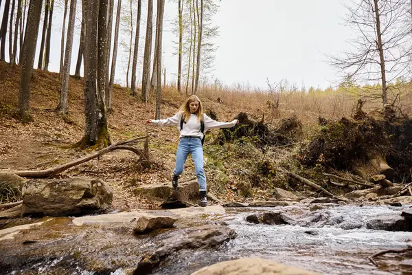 Активная блондинка-туристка в свитере и джинсах пересекает лесной ручей, перепрыгивая через воду — стоковое фото