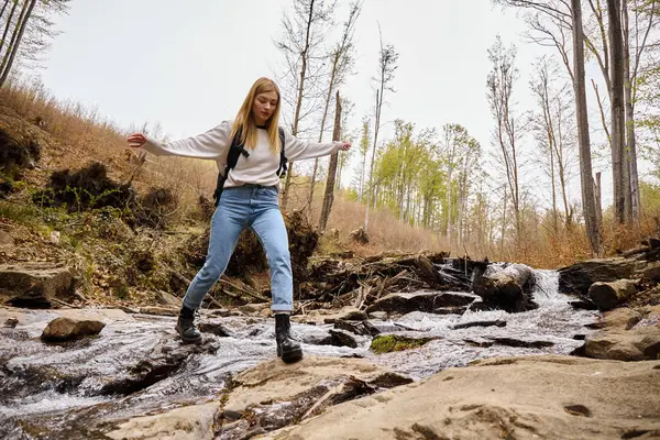 Joyful blonde female hiker wearing sweater and jeans crossing the forest creek jumping over water — Stock Photo