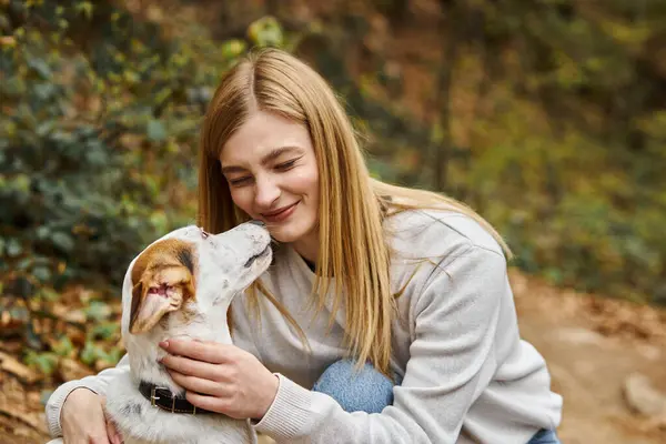 Emocional mujer besando y abrazando a su lindo perro blanco y haciendo cara divertida en el bosque - foto de stock
