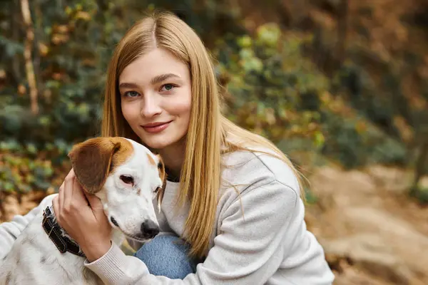 Blonde Frau liebt und umarmt sanft ihren Hund und schaut beim Gassigehen im Wald in die Kamera — Stockfoto