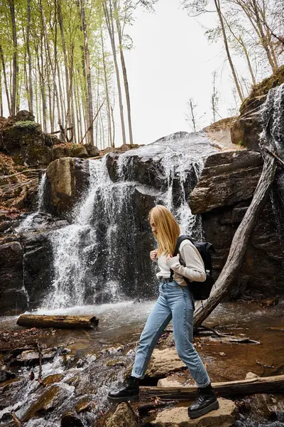 Активная блондинка-туристка, пересекающая лесной ручей, идет по скалам возле водопада — стоковое фото