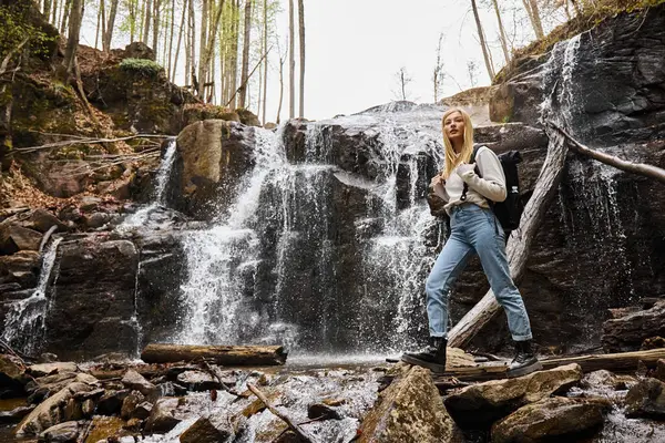 Young blonde female hiker crossing the forest creek walking on rocks near waterfall — Stock Photo