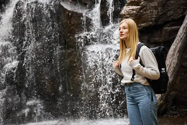 Portrait of young blonde female traveler hiking in beautiful forest and standing near waterfall — Stock Photo