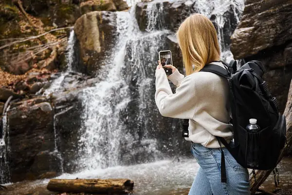 Vista posterior de la joven rubia tomando fotos de la cascada de montaña en el bosque mientras caminaba - foto de stock