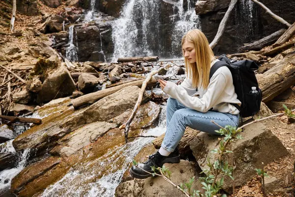 Mujer joven rubia calma con teléfono cerca de la cascada de montaña en el bosque mientras caminaba - foto de stock