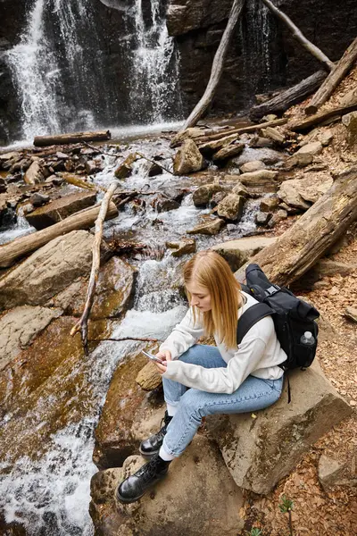 Blonde woman traveler using phone sitting near mountain waterfall in the forest while hiking — Stock Photo