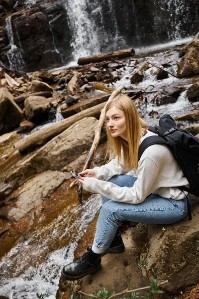 Mujer rubia sosteniendo el teléfono sentado cerca de la cascada de montaña en el bosque mientras caminaba - foto de stock