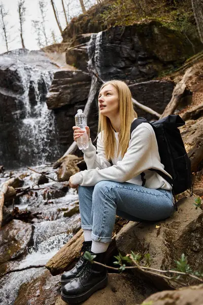 Mujer joven con mochila sentada y sosteniendo una botella de agua y mirando al bosque cerca de la cascada - foto de stock