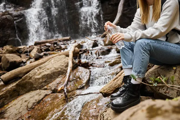 Imagen recortada de mujer en jeans sentada con botella de agua sobre rocas en el bosque cerca de la cascada - foto de stock