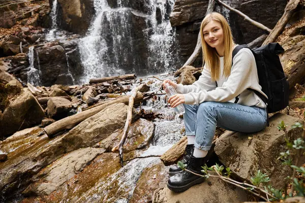 Lächelnde Frau sitzt mit Wasserflasche im Wald in Wasserfallnähe und blickt in die Kamera — Stockfoto