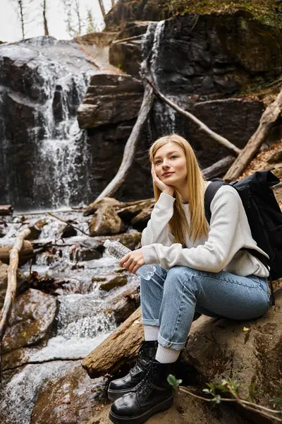 Mujer sonriente sentada con botella de agua y mirando hacia otro lado en el bosque cerca de la cascada - foto de stock