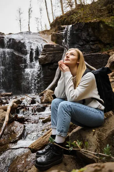 Verträumte Frau sitzt mit Rucksack im Wald in der Nähe des Wasserfalls und blickt in den Himmel — Stockfoto