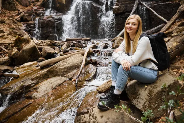 Abenteuerlustige Wanderin sitzt mit Rucksack im Wald am Wasserfall und blickt in die Kamera — Stockfoto