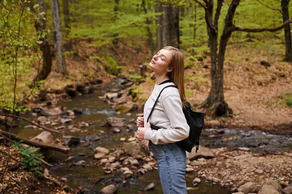 Young blonde female hiker standing with eyes closed in woods. Portrait of relaxed woman in forest — Stock Photo