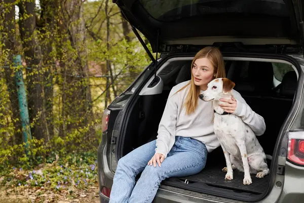 Sorrindo jovem mulher em suéter e jeans abraçando seu cão sentado na parte de trás do carro na floresta em caminhadas — Fotografia de Stock