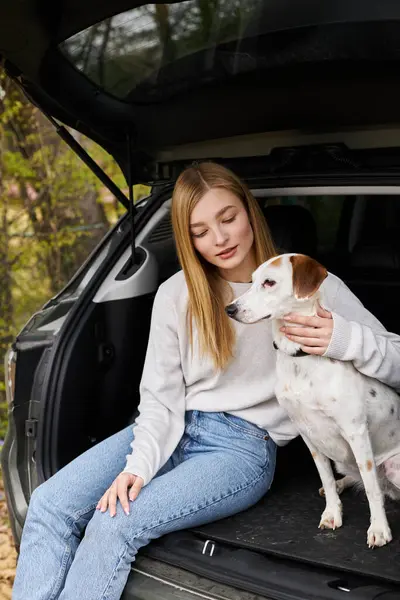 Jovem loira de camisola e jeans abraçando seu cão sentado na parte de trás do carro na floresta em caminhadas — Fotografia de Stock