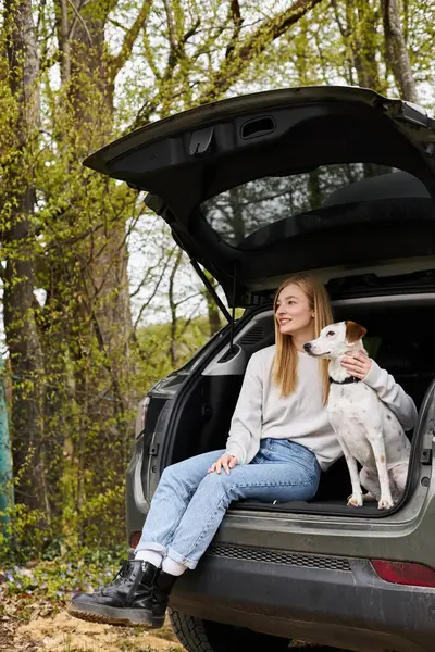 Mujer relajada abrazando a su perro sentado en la parte trasera del coche y mirando hacia otro lado en el bosque en el senderismo - foto de stock