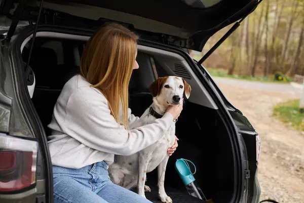 Mujer joven sosteniendo a su perro en las manos y abrazando a su mascota en el descanso de senderismo en la parte posterior de su coche - foto de stock