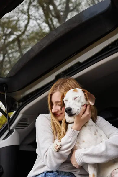 Smiling happy woman hugging dog sitting in back of car in forest at hiking trip halt, eyes closed — Stock Photo