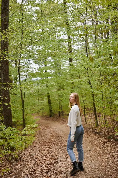 Back view image of curious blonde female traveler hiking in forest in sweater and jeans — Stock Photo