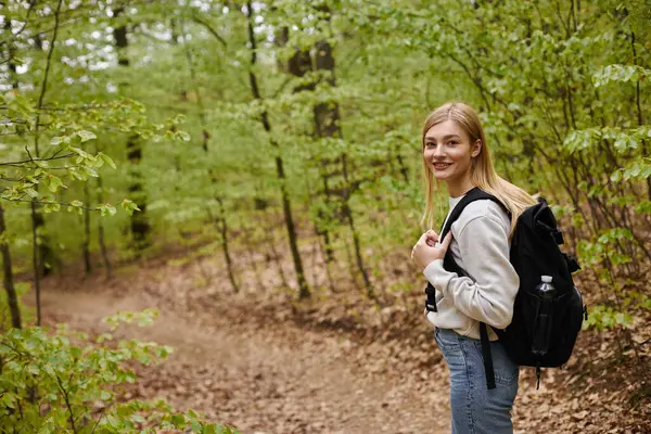 Rückseite Porträt einer lächelnden blonden Reisenden, die im Wald wandert und einen Rucksack trägt — Stockfoto