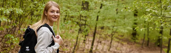 Retrovisore ritratto di una viaggiatrice bionda sorridente che cammina nella foresta e indossa uno zaino, banner — Foto stock