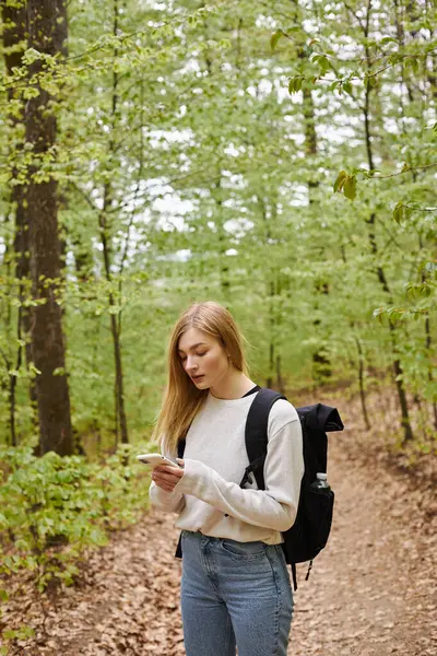 Young blonde female traveler with backpack holding phone hiking in forest and standing on path — Stock Photo