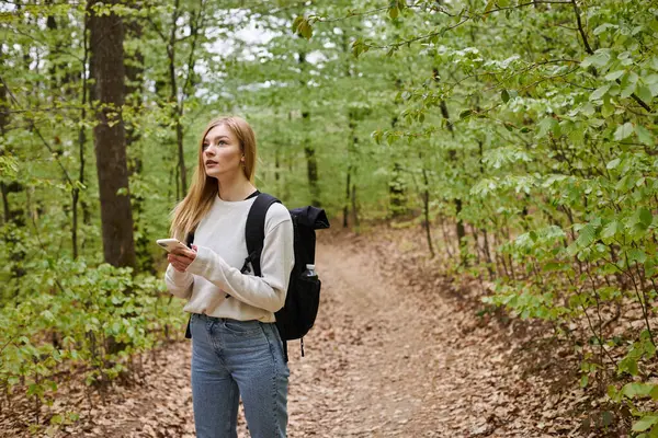 Young blonde female traveler with backpack holding phone hiking in forest looking for direction — Stock Photo