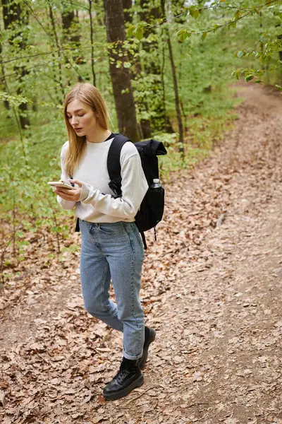 Pretty blonde woman traveler with backpack holding phone walking in forest looking for direction — Stock Photo