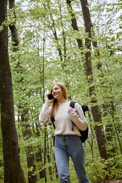 Sonriente mujer rubia viajero con mochila hablando por teléfono caminando en el paisaje del bosque - foto de stock