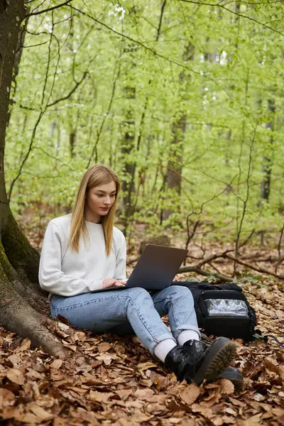 Young female hiker with laptop on her legs wearing sweater, jeans and hiking boots sitting in forest — Stock Photo