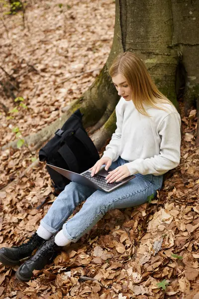 Relajada rubia con portátil en sus piernas trabajando a distancia mientras está sentada en el bosque de viaje - foto de stock